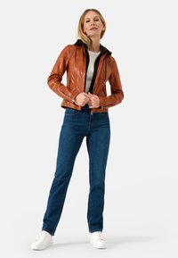 Brun, tætsiddende læderjakke med sort hætte, lynlås foran og to sidelommer, parret med blå jeans og hvide sneakers.