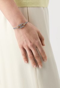 Blauwe leren armband met een gouden accent en een centraal decoratief element, gedragen op een hand tegen een geplooid, lichtgekleurd kledingstuk.