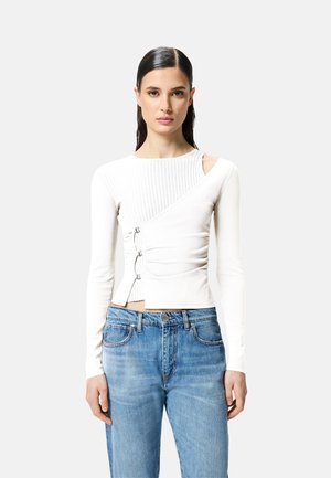 Top lungo bianco a maniche, con texture a coste, design asimmetrico, taglio laterale e dettagli in metallo argentato regolabili. Abbinato a jeans blu.