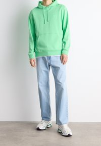 Felpa verde menta con tasca a marsupio, abbinata a jeans dritti blu chiaro e sneaker bianche con accenti verdi e viola.