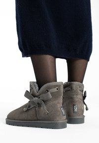Botas de tobillo de ante gris con lazos de cinta, forro suave y suela texturizada. Presentan un parche con el logotipo en la parte trasera.
