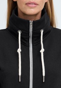Černá mikina na zip s vysokým límcem. Bílé šňůrky se uzlovými konci. Hladká textilie a stříbrné detaily na zipu.