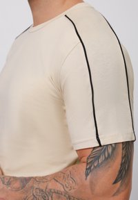 Cremfarbendes T-Shirt mit kurzen Ärmeln, das schwarze Akzente entlang der Nähte aufweist. Der Stoff ist glatt und eng anliegend, wodurch ein tätowierter Arm sichtbar wird.