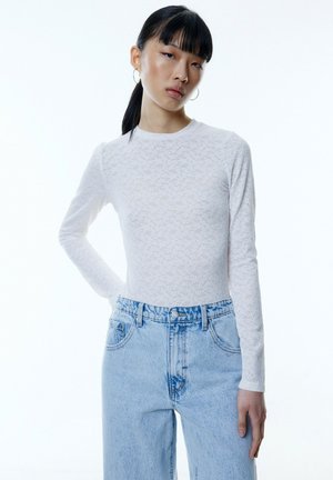 Weißes langärmliges Rippoberteil mit strukturiertem Muster, kombiniert mit hellblauen, hochtaillierten Jeans mit lockerer Passform und sichtbaren Nähten.