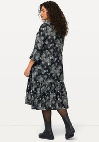 Schwarzes Blumenkleid mit drei Viertelärmeln und einem gestuften Saum, das mit hellgrünen Blumenmustern verziert ist. Kombiniert mit schwarzen Stiefeletten.