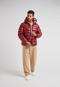 Rød pufferjakke med en skinnende finish, lynlås lukning og en sort-hvid hætteforing. Parret med beige bukser og hvide sneakers.