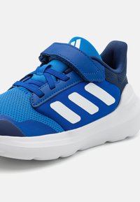 Scarpa sportiva blu con tomaia in rete, strisce bianche, cinturino in velcro e suola ammortizzata. Il design presenta una punta arrotondata e dettagli testurizzati.