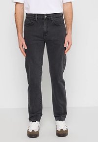 Jean en denim noir à coupe droite, avec poches avant et fermeture par bouton. Porté avec un t-shirt blanc et des baskets.