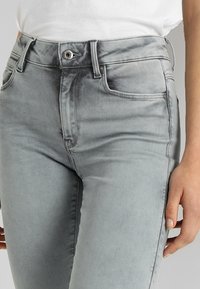 Vaqueros de mezclilla gris con un diseño ajustado, estilo de cinco bolsillos, pasadores para cinturón y un cierre con botón plateado en la cintura. Textura suave.