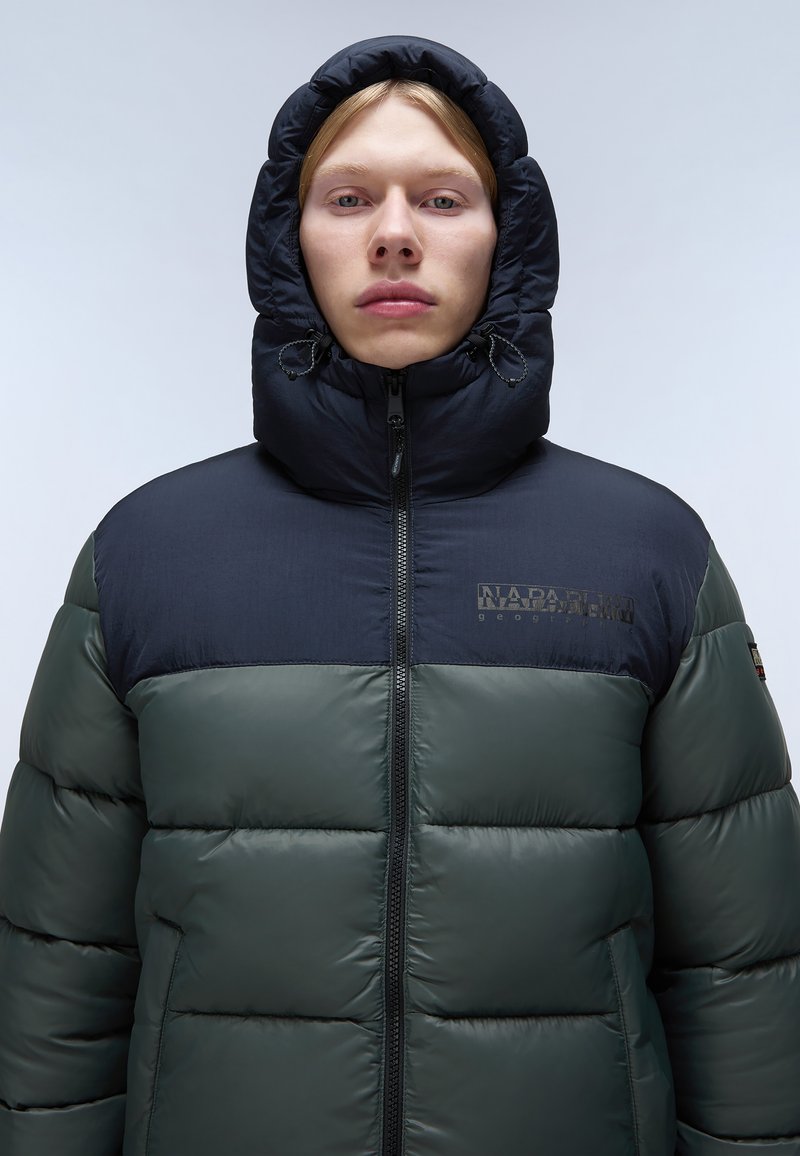 Puffjacke mit einem dunkelgrünen unteren Bereich und einem marineblauen oberen Teil. Sie hat einen hohen Kragen, einen Reißverschluss und ein Logo im Brustbereich.