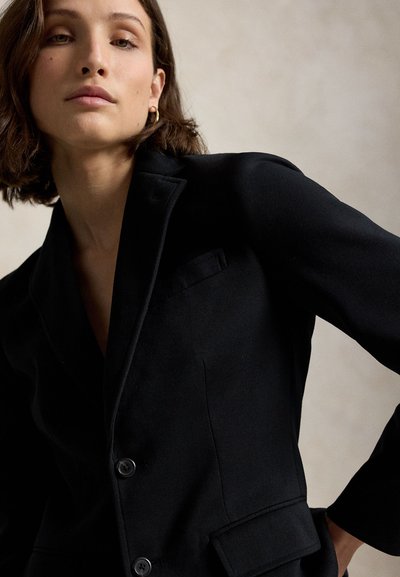 Giovane donna con capelli castani corti che indossa un blazer nero su misura, posa su uno sfondo neutro con un'espressione seria.