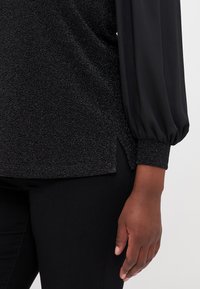 Black top with textured fabric, long silky sleeves, and fitted cuffs. Features side slits at the hem for ease of movement.