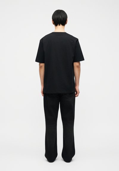 Άτομο με κοντά μαύρα μαλλιά στέκεται με την πλάτη προς τα εμπρός, φορώντας ένα απλό μαύρο κοντομάνικο μπλουζάκι και φαρδιά μαύρα τζιν, μπροστά σε λευκό φόντο.