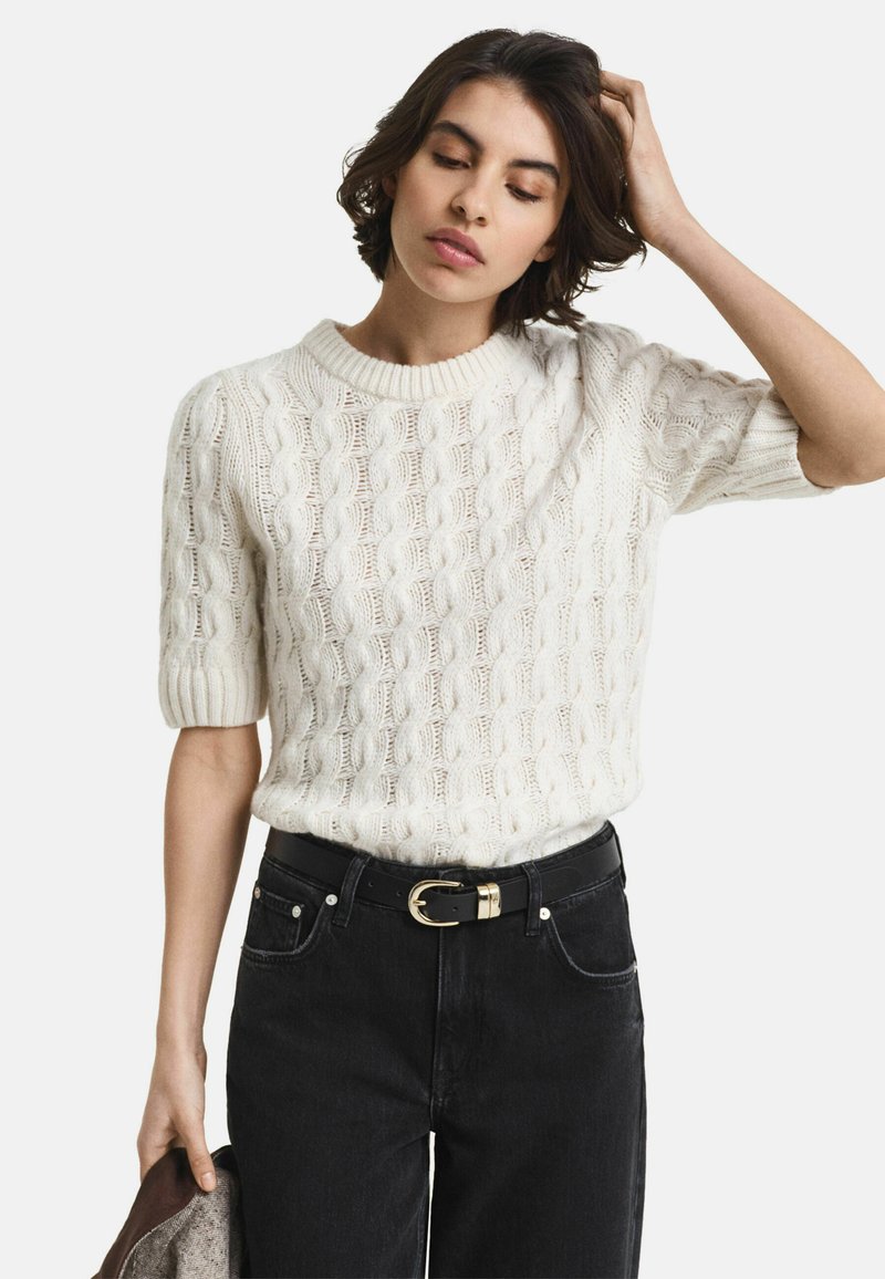 Witte kabelgebreide trui met korte pofmouwen, gecombineerd met zwarte hoge taille jeans en een zwarte riem met een gouden gesp.