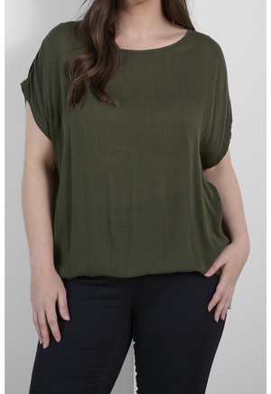 Femme portant un haut ample à manches courtes vert olive et un pantalon noir, debout avec une main dans la poche sur un fond uni.