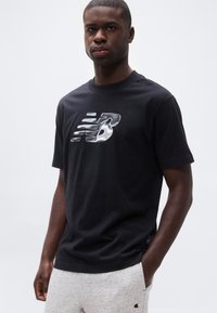 Νεαρός άνδρας που φοράει μαύρο μπλουζάκι με ασημένιο λογότυπο New Balance και ανοιχτό γκρι παντελόνι φόρμας Champion, στέκεται με το ένα χέρι στην τσέπη.