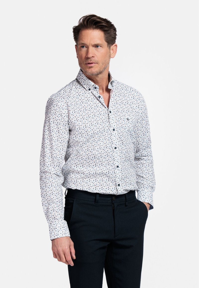 Chemise blanche à manches longues avec un petit motif floral multicolore ; dotée d'un col. Portée avec un pantalon sombre.