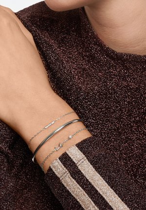 Drie dunne armbanden in zilver- en goudtinten: een elegante metalen cuff, een met kleine heldere stenen, en een met een decoratieve bedel.