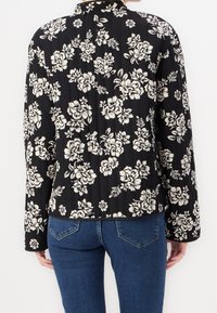 Czarna i biała kurtka w kwiaty z długimi rękawami, o prążkowanej fakturze i wysokim kołnierzu, zestawiona z niebieskimi dżinsami.
