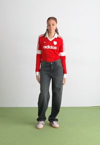 Jersey rojo de adidas de manga larga con detalles en blanco, combinado con jeans oscuros de cintura alta y zapatillas de deporte rosa claro sobre un suelo verde.