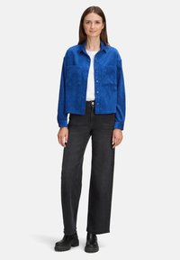 Niebieska kurtka z kordurowego materiału z dwoma przednimi kieszeniami, zapięciem na guziki i luźnym krojem, noszona na białej bluzce i czarnych spodniach typu straight-leg.