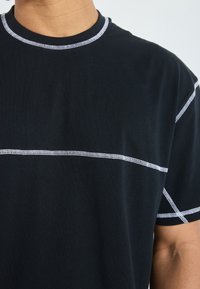 Juoda marškinėliai su apvaliu kaklu, puošti kontrastingais baltais siūlais, sudarančiais geometrinį raštą per krūtinę ir pečius.