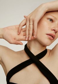Top de tela negra con diseño entrecruzado, que revela el tono de piel. Anillo delicado de oro en los dedos, mostrando la textura y forma natural de las uñas.