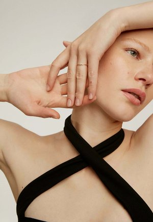 Top de tela negra con diseño entrecruzado, que revela el tono de piel. Anillo delicado de oro en los dedos, mostrando la textura y forma natural de las uñas.