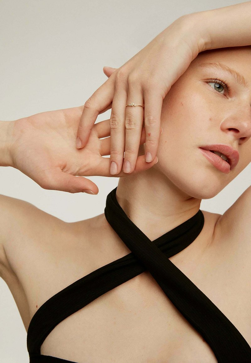 Top de tela negra con diseño entrecruzado, que revela el tono de piel. Anillo delicado de oro en los dedos, mostrando la textura y forma natural de las uñas.