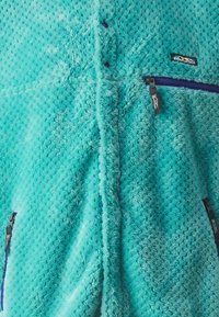 Tyrkise fleece genser med teksturert mønster, med hel glidelås og to sidelommer med glidelås. Liten merkevarelogo på brystet.