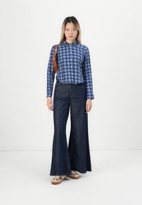 Blå rutete skjorte med knapper, høyt liv mørke denim flare bukser, og lyse sko med et subtilt mønster. Brun skulderveske.