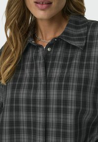 Camisa de cuadros en negro y gris, con cuello de botones y una textura de tela elegante, con un sutil brillo y detalles de hardware en plateado.