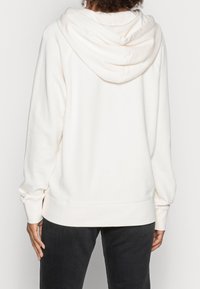 Felpa bianca con un ampio cappuccio, polsini e orlo a costine. Realizzata in un tessuto morbido con una superficie liscia, mostrata di spalle.