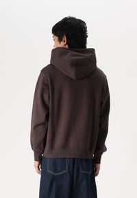 Brown hooded sweatshirt with a relaxed fit, ribbed cuffs and hem, and soft texture. Worn with loose dark denim jeans.
