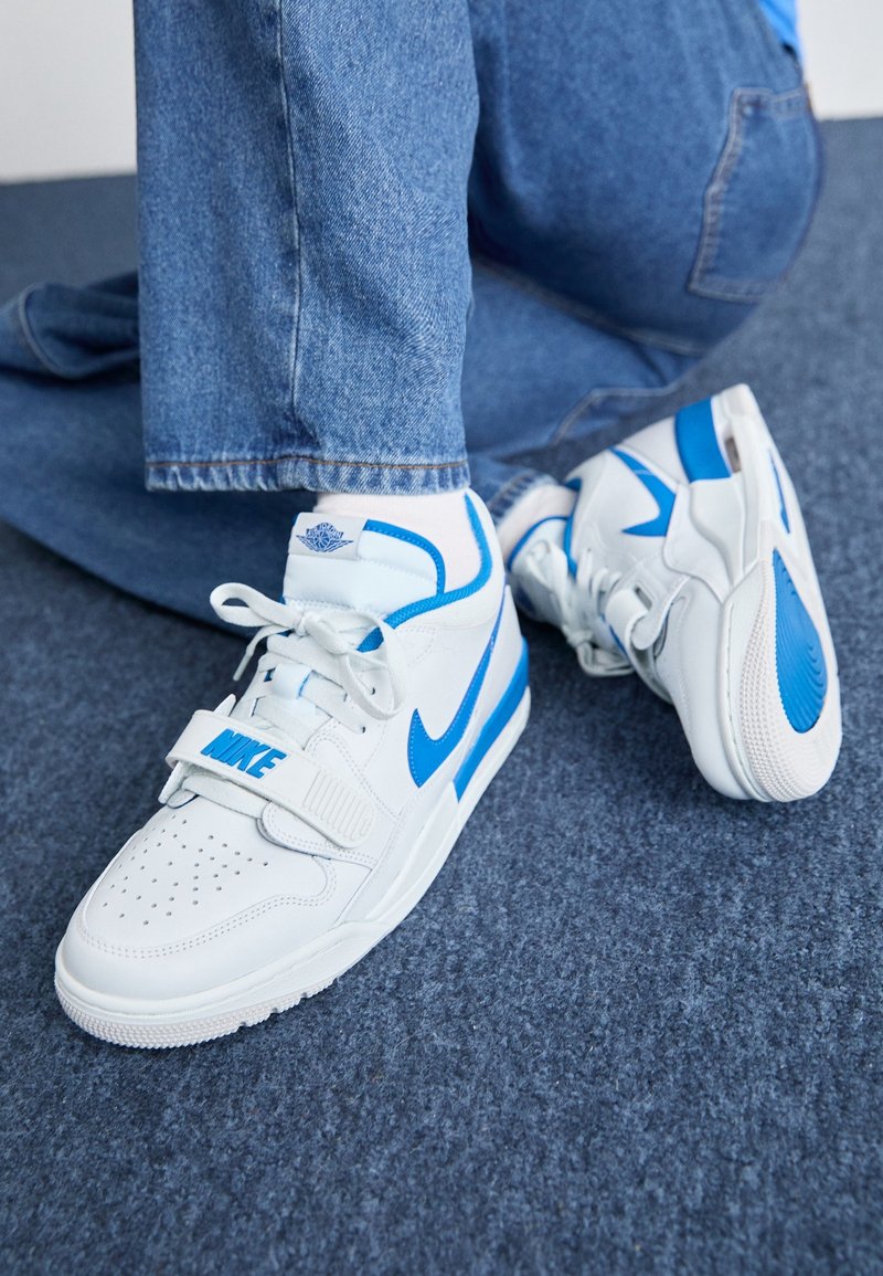 Personne portant des baskets Nike blanches et bleues avec un jean bleu, agenouillée sur un sol recouvert d’un tapis gris.