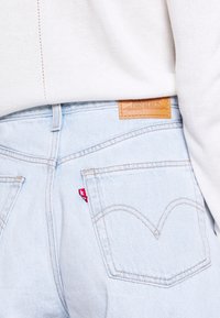 I jeans in denim azzurro chiaro presentano una toppa in pelle marrone sulla vita e un piccolo etichetta rossa vicino alla tasca destra, evidenziando i dettagli di cucitura classici.