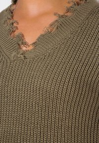 Pull en maille vert olive avec un décolleté en V aux bords effilochés, une surface texturée et une coupe ample.