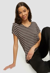 Szary t-shirt w paski z różowymi paskami, z dekoltem w serek i krótkimi rękawami, prezentowany na modelu siedzącym na neutralnym tle.