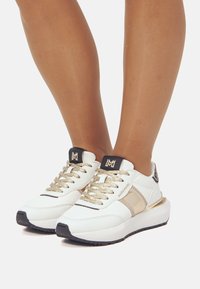 Baskets blanches avec des accents dorés, des lacets texturés et un logo noir sur la languette. Elles présentent une semelle épaisse et une finition lisse.