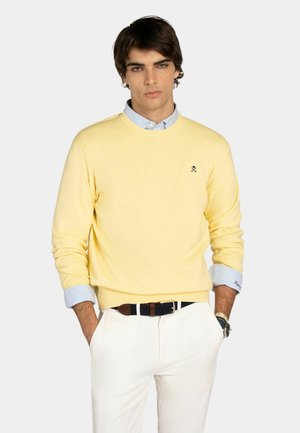Hombre con suéter amarillo de cuello redondo sobre camisa azul clara, pantalones blancos, cinturón negro y reloj de pulsera, de pie con las manos en los bolsillos.
