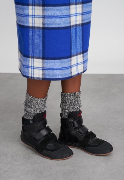 Sorte høyhælte sneakers med borrelås, teksturerte paneler og gummisåler, iført med grå strikkede sokker og et blårutete skjørt.