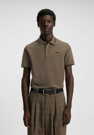 Olivgrön polo med klassisk krage, tre knappar och svart logotyp. Stylad med rutiga, högmidjade, beiga byxor och ett svart bälte.