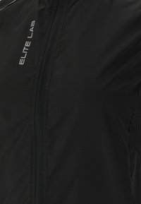 Schwarze Windjacke mit einem Reißverschluss, aus strukturiertem Stoff mit einem bedruckten Logo. Das Design umfasst Mesh-Details an den Seiten.