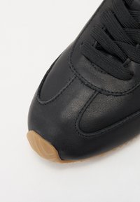 Must musta sneaker tekstuuriga ülaosaga, millel on mustad paelad, siledat pinnat ja beež kummist tallad, millel on kõver muster.