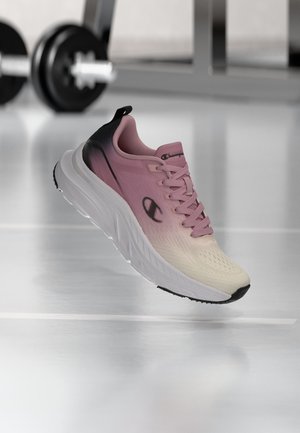 Chaussure de course en dégradé rose et beige avec semelle blanche, logo Champion noir, présentée dans un cadre de salle de sport avec des poids d'exercice flous en arrière-plan.