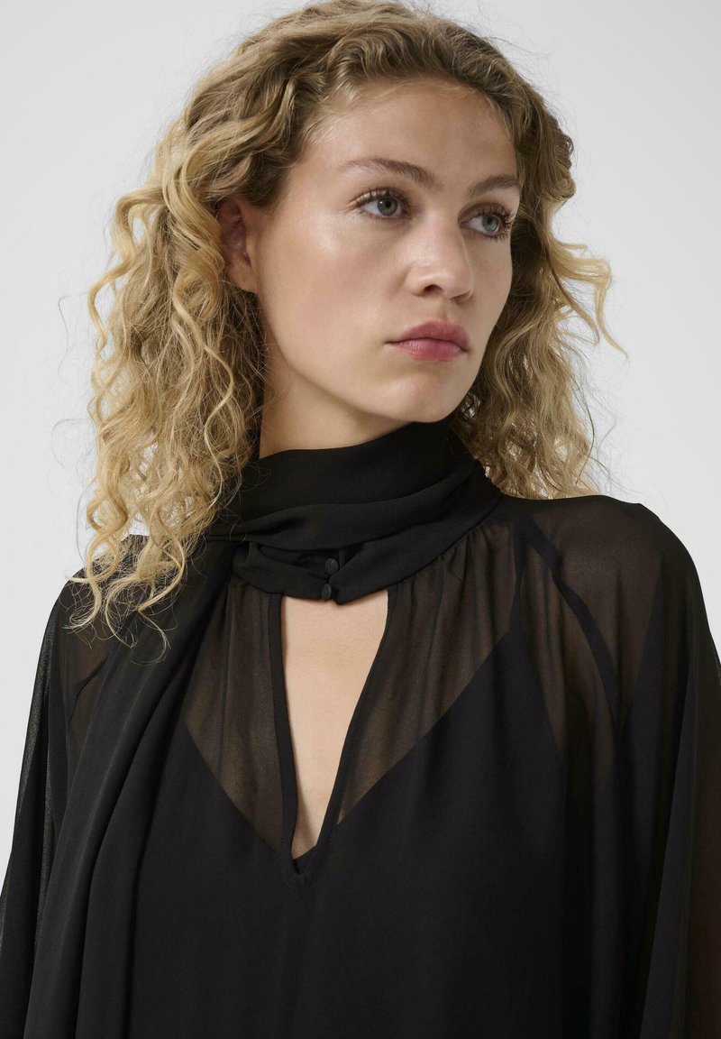 Femme aux cheveux blonds bouclés portant un haut noir transparent avec un col montant et des détails à boutons, regardant sur le côté.