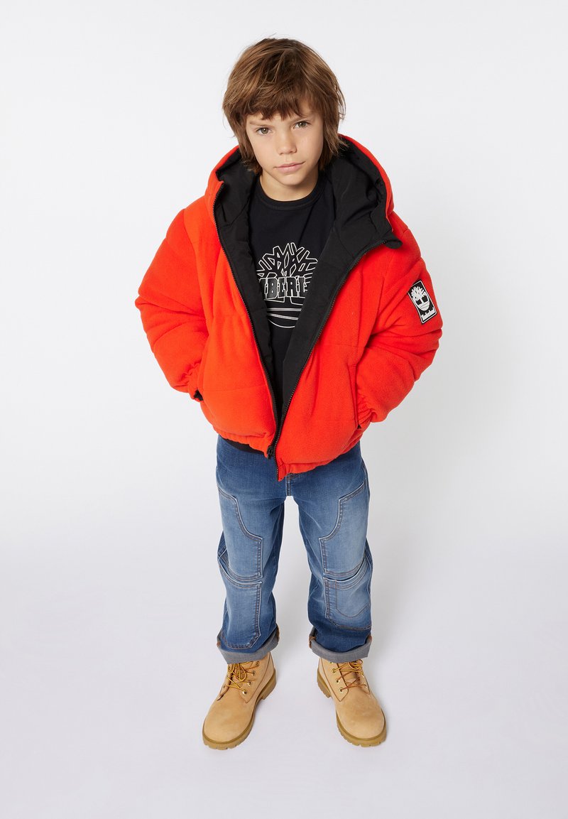 Giacca a vento arancione brillante con cappuccio nero, indossata sopra una maglietta nera con grafica. Jeans blu con cuciture dettagliate e stivali marroni.