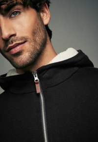 Mustatud kapuutsiga spordijakk, mille kaelus on karvase voodriga, tekstuuriga kangas, hõbedane lukk ja nahkadetail. Suurendatud vaade kaelusele ja näole.