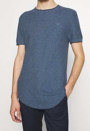 Mężczyzna ubrany w niebieski, krótki rękaw, teksturowany sweter z zaokrąglonym dołem oraz ciemne spodnie w cienkie paski, z ręką w kieszeni.