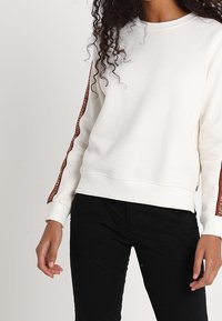Felpa bianca a collo rotondo con strisce marroni texture lungo le maniche, caratterizzata da orlo e polsini a coste, abbinata a pantaloni neri.